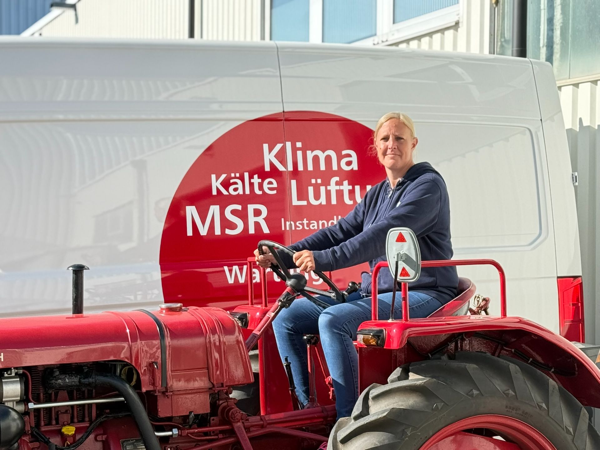 Ein Roter alter Trecker auf dem eine Mitarbeiterin sitzt. Im Hintergrund steht ein Firmenbulli mit Firmenbeschriftung.
